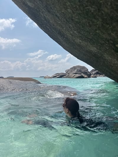 A woman with dark hair pulled back in a bun swims in clear, turquoise water near large rocks. The location appears to be coastal with rocky outcroppings and a partly cloudy sky above. Based on the location indicated, the scene is likely near the coast of Lam Kaen, Thailand, where similar geological features are common.
