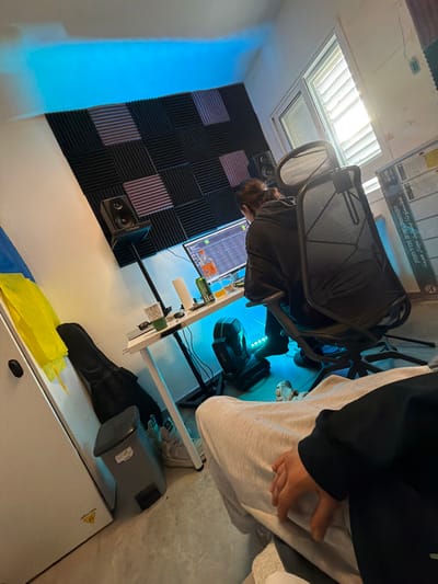 The image, taken in Migdal HaEmeq, Israel, displays an indoor setting consistent with a home music studio. A person with dark hair tied back, wearing a dark hoodie, is seated with their back towards the viewer in a black mesh office chair, facing a computer monitor that displays digital audio workstation (DAW) software.
On the white desk in front of the monitor are a green Hever soda can, a clear glass containing a beverage, a roll of white paper towels, a white paper cup, and an orange bottle. A blue-green LED light strip illuminates the desk's underside. Two studio monitor speakers, one identified as a Kali Audio LP-6 V2, are positioned on stands flanking the monitor area. The wall behind the desk is covered with a grid of black and purple acoustic foam panels. Blue LED strip lighting is affixed to the ceiling corner directly above these panels. Natural light enters through a window with white horizontal blinds on the right side of the frame.
In the immediate foreground, the lower torso and right hand of a second individual are visible. This person is wearing light gray sweatpants and a dark long-sleeved garment, with their right hand resting on their thigh.
To the left of the desk, a white wardrobe or cabinet has a yellow fabric draped over its top corner. A black, soft-sided instrument case, likely for a guitar, leans against the wardrobe. A gray, pedal-operated trash can is positioned adjacent to the instrument case. A pair of white sneakers is visible on the floor near the desk. A large, upright cardboard box with Hebrew text and an image of a speaker, likely packaging for audio equipment, stands partially visible on the far right near the window. The floor is a light-colored, smooth surface.