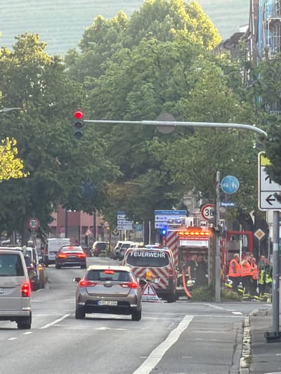 The image depicts a street scene in Singen (Hohentwiel), Germany, where a fire truck is present, indicating an emergency response. The fire truck, with its lights illuminated, is positioned on the right side of the road. Several individuals in high-visibility vests, likely firefighters and emergency personnel, are seen near the truck. The traffic light in the foreground is red. Several cars are on the road, stopped or moving slowly, suggesting traffic disruption due to the emergency. The environment includes trees lining the street and buildings in the background. Road signs, including a speed limit sign (30), are visible.