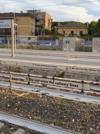 The image depicts a railway scene in Ladispoli, Italy, characterized by multiple warnings against crossing the tracks. In the immediate foreground, two parallel railway tracks are visible, embedded in a bed of grey and light-colored ballast stones. Immediately above and between the tracks are horizontal white barriers with red diagonal stripes, indicating a restricted area. Attached to these barriers are white rectangular signs featuring black text; one prominent sign clearly reads "Do not cross the railway lines" accompanied by a red circular icon depicting a stylized person with a diagonal line through it, signifying prohibition.

Above the railway lines and barriers, a concrete train platform extends horizontally across the midground. The platform surface is light grey with tactile yellow warning strips along its edge. On the vertical face of the platform, another white sign with black text is visible, stating "Vietato attraversare i binari" (Italian for "Do not cross the railway lines") and partially revealing its English translation alongside a "no pedestrian" icon. A slender, light-colored pole, possibly a light fixture or signal support, stands on the platform to the left. Behind the platform, a metal fence separates the railway area from an adjacent space where several cars are parked, including a silver hatchback, a dark sedan, and a white vehicle.

The background features several residential buildings under a partly cloudy sky. To the left, a multi-story building with beige brick cladding and a dark roof is visible, with multiple television antennas on its rooftop. In the central-right background, a distinct, single-story house with yellow-ochre walls and a reddish-brown tiled roof featuring several decorative chimneys or vents is prominent, partially obscured by green trees and foliage. Other buildings and dense greenery are present further in the background. The sky is a light blue with scattered white clouds, indicating daylight conditions. No individuals are present in the frame.