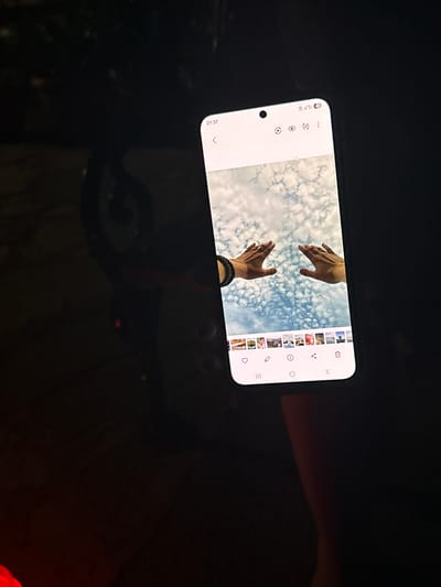 A smartphone is held in the dark, displaying a photo of two hands reaching up towards a blue sky filled with white clouds. The phone screen also shows a gallery of smaller thumbnail images at the bottom, a navigation bar, and status icons at the top. The background of the image is dark and out of focus, with hints of red light visible. It is impossible to determine the location within Vrnjacka Banja Municipality, Serbia from the provided image.