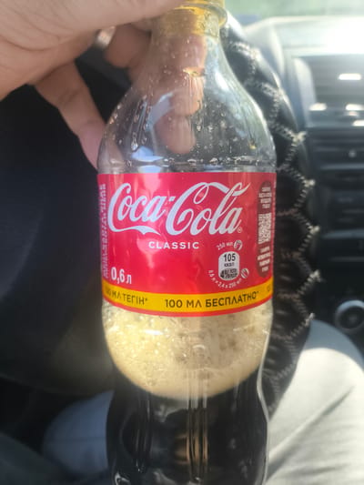 A person's hand holds a clear plastic bottle of Coca-Cola Classic. The bottle is partially full of a dark brown liquid with a foamy head. The label is red and white, with the Coca-Cola logo prominently displayed. Text on the label indicates "0.6 L", "250 ML", "CLASSIC", and nutritional information. A yellow band below the red label contains more text. The background appears to be the interior of a car, with a black leather steering wheel visible. The scene is brightly lit, suggesting it is daytime. There is no information available about the location being Abay, Kazakhstan.