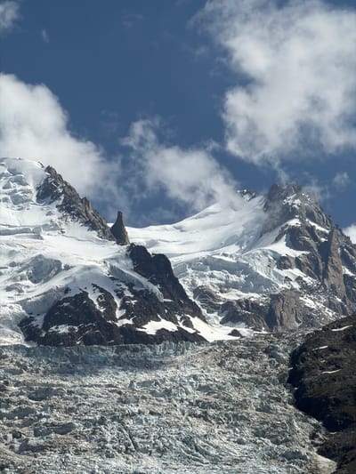 The image shows a mountain range covered in snow and ice, with a glacier in the foreground. The sky is blue with scattered clouds. The mountains are rugged and steep, with rocky outcrops and patches of snow. The glacier appears to be a large, broken mass of ice, with crevasses and debris visible. The scene is typical of the Alps, and likely depicts a view of Mont Blanc from Chamonix-Mont-Blanc, France. No people or artificial objects are visible in the image.