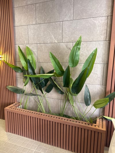 The image shows a close-up, eye-level view of a modern planter box filled with artificial greenery, positioned against a tiled wall. The planter box is constructed from vertical, reddish-brown wooden slats. The greenery consists of several large, elongated, dark green leaves with prominent parallel veins, arranged in a lush display. Some of the leaves are a lighter, more vibrant green, creating a two-toned effect. The wall behind the plants is covered in rectangular, light gray tiles with subtle veining. To the left of the planter, a wooden slatted panel is visible, with a light source emanating from the top left corner, casting a warm glow. The floor beneath the planter is tiled with square, beige tiles. The overall impression is one of contemporary interior design. The scene is a generic interior space, and no specific location in Ayn al Basha, Jordan, can be inferred from the visual information provided.