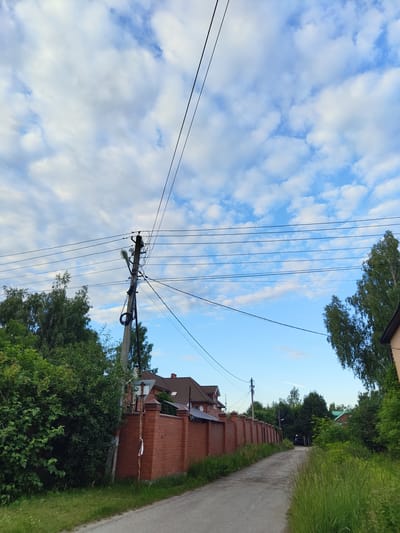 This media file depicts a roadside scene in Tsibino, Russia. The image is taken in a vertical orientation, capturing a road on the right side of the frame, bordered by lush green grass and trees. Overhead, a clear blue sky with scattered white clouds dominates the upper portion of the image. A power pole with multiple wires stretches across the frame, with a street lamp attached to it.
To the left of the road and behind a prominent brick wall, several houses with tiled roofs are visible, nestled amongst dense green foliage. The overall setting suggests a semi-rural or suburban environment. The time of day appears to be late afternoon or early evening, indicated by the soft lighting and the clear sky. There is no activity or event clearly taking place, other than the presence of a parked car in the distance down the road. No text is visible in the media file.