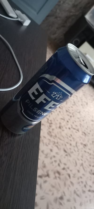 A blue and silver Efes Pilsener beer can is shown tilted on its side, resting on a dark brown wooden surface. The can's tab is open. The background consists of a textured, light-colored surface, possibly a wall or floor. A white electrical cord and plug are partially visible in the upper left corner, also on the wooden surface. The scene appears to be indoors, in Menzelinsk, Russia, possibly on a table or counter.