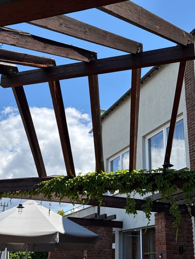 An outdoor scene beneath a wooden pergola covered in green ivy. The pergola's dark wooden beams are visible against a bright blue sky with scattered white clouds. Beyond the pergola, a light-colored building with several white-framed windows and a red brick chimney is visible. In the foreground, a portion of a large, light grey umbrella is present. This image was taken in Duszniki, Poland.