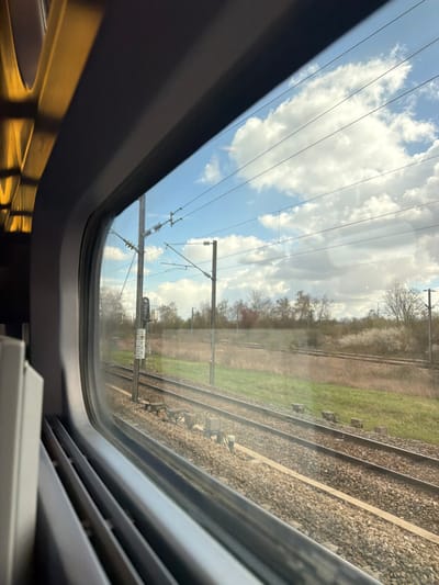 The image displays a view from inside a train, looking out through a rectangular window. The scene depicts a railway environment within the city of Chelles, France.
**Environment:**
* **Foreground and Midground:** Multiple parallel railway tracks are visible, constructed with steel rails on gravel ballast. Small, rectangular concrete blocks are positioned alongside the tracks. A strip of green grass and dry, brown vegetation runs adjacent to the railway line.
* **Background:** A dense line of deciduous trees forms the horizon, exhibiting varying stages of early spring foliage, including some with reddish leaves, nascent green growth, and white blossoms.
* **Sky:** A partially cloudy blue sky with scattered white cumulus clouds is overhead.
* **Infrastructure:** Overhead electrical catenary lines, characteristic of electrified railway systems, traverse the sky. These lines are supported by at least two robust utility poles. One pole, located in the left midground, features a vertical apparatus with visible markings, possibly a signaling or measurement device.
**Lighting and Conditions:**
* The scene is illuminated by natural daylight, suggesting fair weather. Clear shadows are cast on the ballast and ground.
**People, Subjects, Objects, Actions, Interactions:**
* No people are present or discernible in the image.
* No specific actions or interactions are depicted.
* The primary subjects are the railway infrastructure and natural landscape.
* Objects include the train window frame, railway tracks, gravel ballast, concrete blocks, utility poles, catenary lines, and trees.