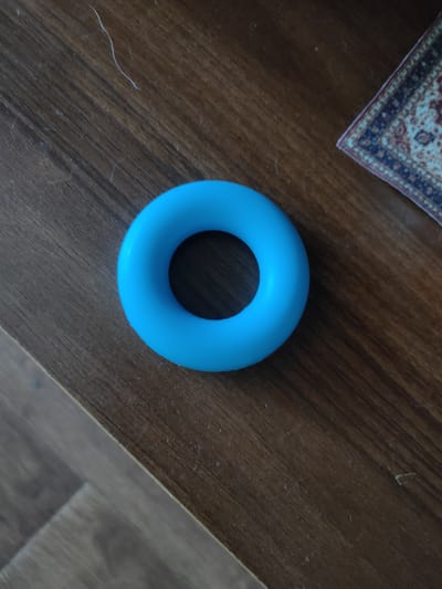 A bright blue, circular object, possibly a hand exerciser or stress toy, rests on a dark brown wooden surface. The wood grain is clearly visible, with some lighter streaks and dust particles scattered across it, suggesting it might be a table or desk. In the upper right corner, a corner of a small rug or tapestry with a traditional pattern in red, blue, and cream is visible. The background is out of focus, but the lighting suggests it is daytime, possibly indoors. No people are visible, and no text is present. The overall impression is a simple, still life composition. The location context provided is Salavat, Russia.