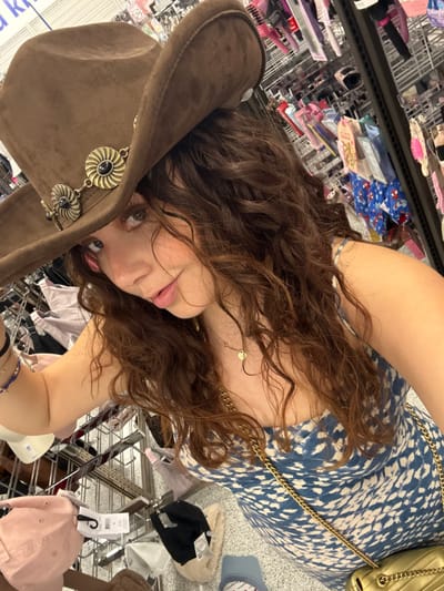 A woman with curly brown hair and a brown cowboy hat with circular adornments poses for a selfie in a retail environment. She wears a blue and white printed dress with a gold chain purse strap visible. Racks of merchandise, including hair accessories and clothing, surround her, creating a cluttered background. Her face is slightly tilted, and she looks toward the camera.
