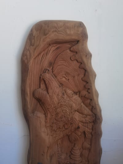 This is a close-up, vertical shot of a wooden carving, likely a decorative wall hanging, set against a plain white wall. The carving depicts a wolf howling at a moon, rendered in a stylized, relief style. The wood itself has a prominent grain pattern, with variations in color and texture that add depth to the artwork.
The wolf's head is the central focus, with its mouth open in a howl. The fur is textured with intricate carving lines, suggesting movement and detail. Above the wolf, a crescent moon is carved, with a jagged, mountainous landscape or rocky terrain surrounding it. The edge of the carving follows the natural, irregular shape of the wood slab, giving it an organic and rustic feel. Below the wolf's head, there are carved shapes that appear to represent pine trees or other foliage, suggesting a forest or wilderness setting.
The lighting in the image is diffused, casting soft shadows that highlight the carved details. There is no visible text in the image. The scene evokes a sense of the wild, perhaps a clear night in a remote, natural environment. The overall impression is one of rustic artistry and a connection to nature. The location context is Cherkessk, Russia, but this information is not directly discernible from the visual cues in the image itself.