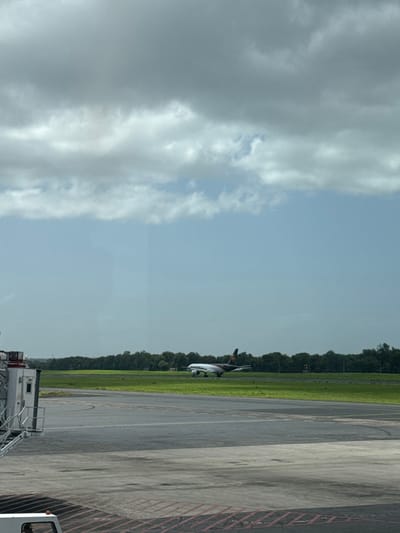 The image displays an outdoor scene at what appears to be an airport or airfield, viewed under a partly cloudy sky during daylight hours. The perspective is looking out over an expanse of tarmac and grass towards a line of trees and an aircraft.

The sky occupies the upper portion of the frame, showing a dynamic mix of light blue patches and significant cloud cover. On the left side, large, thick cumulus clouds, varying in shades from bright white to soft grey, dominate the sky. Towards the right, the sky is clearer, revealing a lighter blue hue with fewer, more scattered clouds.

A distinct horizontal line of dark green, densely packed trees stretches across the middle of the image, forming the horizon. Below the treeline, there is a vibrant strip of green grass.

Positioned on the right side of the frame, partially against the grass and partially above the tarmac, is an aircraft. It appears to be a small to medium-sized jet, possibly a regional or private aircraft. Its fuselage is light-colored, likely silver or white, while its tail and engine areas appear darker. The aircraft is mostly visible from its side, suggesting it is parked or slowly taxiing in the mid-ground.

The lower half of the image is dominated by a grey asphalt or concrete tarmac surface, which shows some textural variations and darker sections. Faint red-orange markings are visible along the bottom edge of the tarmac on the right side.

In the bottom right corner, a portion of a white ground support vehicle or structure is visible. It includes a set of stairs, suggesting it might be an air stair unit or a mobile boarding ramp, indicating proximity to an aircraft or terminal area.

The overall scene suggests a typical airport environment with an aircraft either preparing for departure, having recently arrived, or simply parked on the airfield. The lighting indicates it is daytime, with a bright, but not intensely sunny, atmosphere due to the cloud cover. No specific human activity or discernible text is visible within the frame.