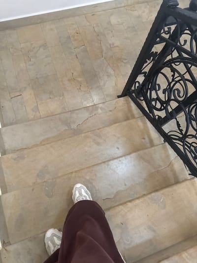 The image presents an overhead, first-person view looking down a worn indoor staircase. The lower legs and feet of an individual are visible, positioned on one of the upper steps, clad in white athletic shoes with gray accents and loose-fitting dark brown or maroon trousers. The staircase is constructed from light-colored, possibly beige, stone or concrete, exhibiting extensive signs of wear, including widespread discoloration, surface cracks, and flaking material on both the treads and risers. Above the visible steps, a landing surfaced with similar light-colored rectangular tiles is present, showing comparable signs of age and degradation, with a plain white baseboard running along its left edge. A section of a black, ornate wrought-iron railing, featuring intricate scrollwork and geometric patterns, is visible along the right side of the staircase. The scene depicts an interior architectural space. The image itself provides no visual identifiers to confirm a specific location within the city of Aragats, Armenia.
