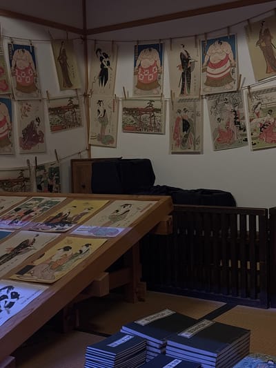 The image depicts an indoor scene within the Sumida Hokusai Museum in Sumida, Japan, showcasing a collection of traditional Japanese ukiyo-e prints. The prints, featuring diverse subjects such as sumo wrestlers, geishas, and landscapes, are displayed on a wall, hung with clothespins on a string, and also laid out on angled wooden display stands. Several stacks of books with Japanese characters on their covers are visible on the floor in the foreground. The environment is dimly lit, with warm ambient light highlighting the artworks and the wooden furniture. A dark fabric is draped over a piece of furniture in the background, partially obscuring it. The overall impression is one of an art exhibition, with an emphasis on historical Japanese art.