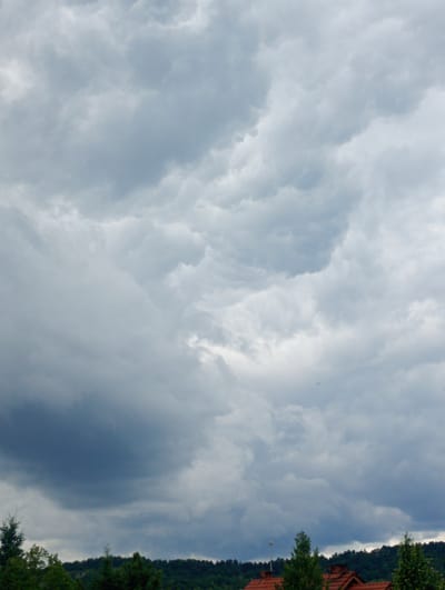The image captures a cloudy sky above a landscape of trees and rooftops in Nowa Wieś Szlachecka, Poland. The sky is filled with dramatic, grey clouds, suggesting overcast weather, possibly a storm brewing. In the distance, a line of dark green trees forms a dense treeline across a hill. Below the trees, glimpses of red-tiled rooftops indicate a residential area. A small, dark object, possibly an airplane, is visible in the upper right portion of the sky. The overall impression is a moody, natural scene with human habitation present in the background.