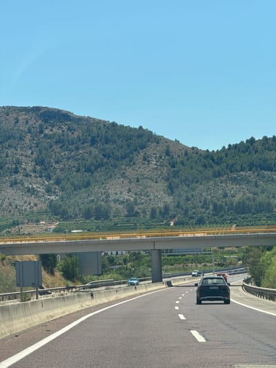 An outdoor, eye-level, medium shot captures a clear blue sky above a mountainous landscape with scattered green trees and some terraced fields. A yellow overpass stretches horizontally across the mid-ground. Below the overpass, a highway curves to the right, with a black car in the foreground and several other vehicles visible further down the road. A concrete barrier with metal railings lines the highway. The location is identified as being on the highway AP-7 near Canals, Spain.