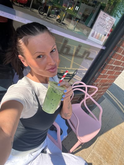 A self-portrait captures a woman sitting outdoors on a bright, sunny day, holding a green beverage. The woman, with dark hair and dark eyes, looks directly at the camera, exhibiting a calm expression. She wears a white short-sleeved top beneath a dark ribbed tank top. Her fingernails are painted a light blue, and she has visible tattoos on her left arm and shoulder. She holds a clear plastic cup containing a vibrant green, ice-filled drink, likely a matcha latte or smoothie, with a red and white striped straw.
The setting appears to be an outdoor seating area of a cafe or shop. A light pink, modern-style metal chair is positioned beside her, with a blue object partially visible further down. The ground is a light-colored paved surface, featuring tactile warning strips. In the background, a brick building facade is visible with a large window. Reflections in the window reveal an outdoor street scene, including greenery, what appears to be parked vehicles, and other urban elements.
Several pieces of text are visible on a sign displayed in the shop window: "AWARD WINNING FRESHLY BLENDED SMOOTHIE" and "Sold Here", accompanied by illustrations of various smoothies. Part of what appears to be the shop's name, "biin", is also reflected in the window. Another reflected poster shows text that includes "GHOST HILL 1-2023". Strong sunlight casts distinct shadows, indicating it is daytime. The image conveys a relaxed moment at an outdoor establishment in Waverley, United Kingdom.
