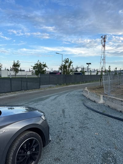 This image captures a semi-developed outdoor area in Costinești, Romania, under a partly cloudy blue sky. The foreground on the left shows the front quarter panel and wheel of a dark gray Skoda vehicle, identified by a "MONTE CARLO" badge on the fender, parked on a light gray gravel surface. This gravel extends across the lower half of the image, forming a curved path or lot.
To the right, a black flexible hose or cable lies along the edge of the gravel, running alongside a series of stacked, weathered concrete Jersey barriers. Beyond these barriers, a chain-link fence is visible, separating the gravel area from an unkempt patch of ground.
In the midground, a dark mesh privacy fence, supported by metal posts, stretches horizontally across the scene. Behind this fence, several young deciduous trees with green foliage are planted in a row. Beyond the trees and fence, multiple white, rectangular modular units or containers are arranged. Further into the background on the right, a tall red and white lattice communication tower stands prominently against the sky, equipped with multiple antennas. To the left of the tower, a solar panel array mounted on a pole is visible, and further left, a modern street light on a tall pole. Distant structures, including large metal framework installations (possibly part of an event stage or amusement park structure) and blurred buildings, are discernible against the horizon, indicating an urban or semi-urban environment consistent with a resort town. No people are visible in the scene.