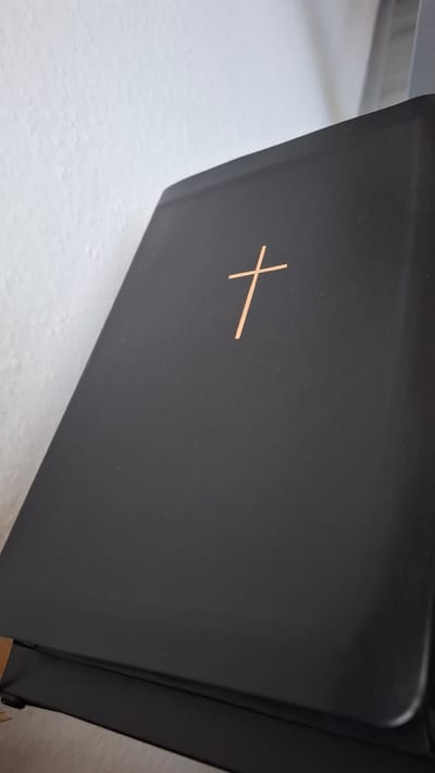 The image shows a close-up, eye-level view of a dark, possibly black, book with a copper-colored cross embossed on its cover. The book is positioned at an angle, with its spine facing towards the right side of the frame. The cover appears to be made of a smooth, matte material. The cross is centrally located on the visible surface of the cover. To the left of the book, a light-colored, textured surface, possibly a table or wall, is visible, indicating a simple indoor setting. The lighting is diffused, suggesting an indoor environment without direct sunlight. No people are visible in the image. The image focuses solely on the book and its decorative cross, with no discernible text present. The overall impression is one of simplicity and religious symbolism.