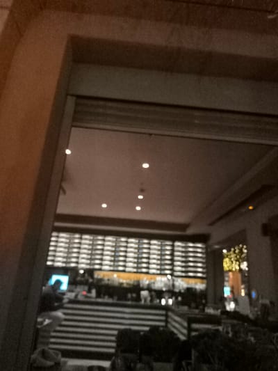The image captures a dimly lit indoor scene, looking into a commercial establishment, likely a bar or restaurant, through a dark, architectural opening or doorway. The framing on the left and top suggests an archway or a thick wall structure.

Inside, the ceiling features several small, bright recessed lights, illuminating the space. A dominant feature is a large, grid-like wall display in the background, appearing to be a well-stocked wine or liquor rack with numerous bottles, many of which are illuminated from behind or below, creating a luminous pattern. Below this display is a bar counter, where various bottles and glassware are faintly visible.

On the left side of the bar, a single person is seen, partially obscured, appearing to be seated or standing. A bright blue screen, possibly from a laptop or tablet, is prominently visible in front of them. In the foreground, there are dark, possibly striped structures that could be banquettes, planters, or partitions. To the far right, some out-of-focus, warm-toned lights, resembling decorative string lights, add a festive touch.

The setting suggests evening or nighttime due to the reliance on artificial lighting. No specific weather conditions are discernible. There are no explicit location cues visible in the image itself to confirm Palaio Faliro, Greece, apart from the provided context. No emotions are discernible, and there is no visible text.