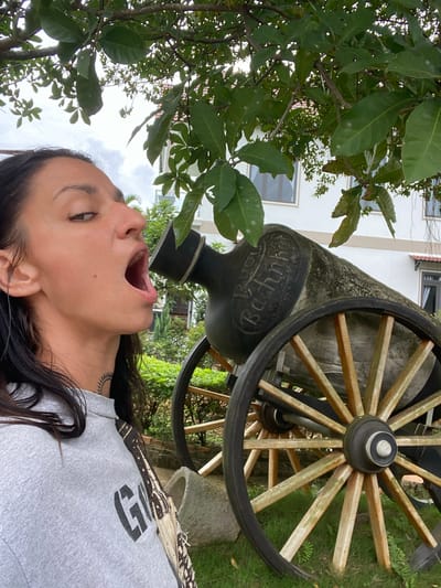 In Thôn 2, Vietnam, a woman with dark hair and a sun tattoo on her neck is posing with an antique cannon. She is wearing a gray sweatshirt with the word "GOOD" printed in large black letters on the front. The cannon, made of stone and wood, has a visible inscription on its barrel that appears to read "Buhlmann." The scene is outdoors, with lush green foliage and a white building in the background. The woman has her mouth open, as if interacting playfully with the cannon.
