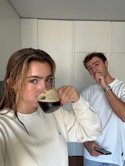 An indoor photograph captures two young adults in what appears to be a residential setting. In the foreground, a female with wet, light brown hair and a cream-colored knitted GANT sweater drinks from a clear glass mug filled with a dark, foamy beverage. Her left hand, adorned with red nail polish, holds the mug. Slightly behind her and to the right, a male in a white t-shirt and light-colored pants poses with his right hand covering his chin and mouth. He wears a silver-toned watch on his left wrist, which also shows a dark tattoo or mark on the forearm, and holds a dark smartphone in his left hand. Both individuals are looking towards the camera. The background consists of light-colored, flat-panel cabinet or wardrobe doors. The specific location within Cidade da Maia, Portugal, is not discernible from the visual information present in the image.