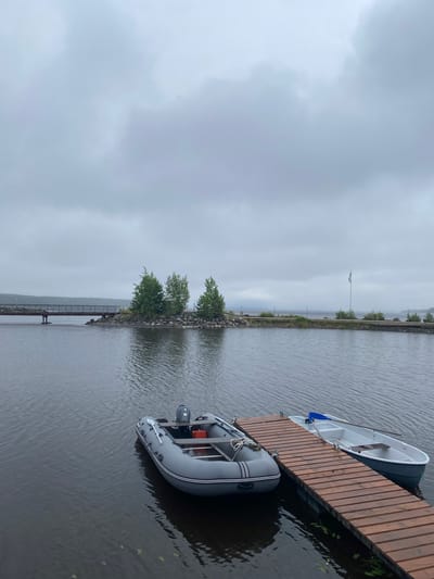 The image depicts a tranquil scene on the water, likely in Kirkkolakhti, Russia, under a cloudy sky. In the foreground, two boats are moored at a wooden dock. The boat closer to the viewer is a grey inflatable dinghy with a small outboard motor and an orange container visible inside. A paddle rests on its side. The second boat, a white rowboat with blue oars, is tied to the dock. The water is calm and dark, with ripples reflecting the overcast sky.
In the mid-ground, a small, rocky island is dotted with three evergreen trees. A pedestrian bridge, made of metal, extends from the left side of the frame towards the island. Beyond the island, the water stretches out to a distant shoreline, also under the gray, diffused light of the clouds. A flag on a pole is visible on the right side of the mid-ground, indicating a populated area or a specific landmark. The overall atmosphere is subdued and peaceful, suggesting a quiet, perhaps misty, day.