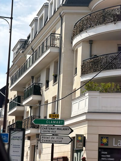 The image depicts a street view in Châtenay-Malabry, France, with a focus on the facade of a modern building and various signs. The building features multiple balconies with black metal railings and is adorned with planters containing dried foliage. The architecture is characterized by light-colored stone cladding and white trim.
Several street signs are prominently displayed. One green sign reads "CLAMART" with an arrow pointing to the left, and another sign below it indicates "CHÂTENAY-MALABRY -CENTRE-VILLE". Adjacent to these are signs for "Hôtel de Ville" and "Commissariat". A yellow sign with "D 986" is also visible.
Further down, a shop sign for "La Vie Claire" is seen, advertising "ÉPICERIE", "PRODUITS FRAIS", and "FRUITS ET LÉGUMES". Below this, a sign for "SAM'NET PROPRETE" lists services like "Nettoyage résidences", "Bureaux", "Commerces", and "Service". A blue sign with a bicycle symbol is also present.
The overall scene suggests a bright day, likely in the daytime, given the sunlight casting shadows on the building. The perspective is angled, looking up at the building from a street level. The presence of street signs and shop names strongly indicates an urban or suburban commercial area. No people are clearly identifiable in the image, though a glimpse of someone in dark clothing is visible near the "La Vie Claire" sign. The weather appears clear with some sky visible between the building structures.