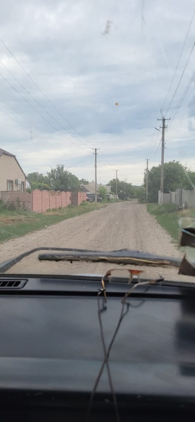 The image shows a view from inside a vehicle looking through the windshield. The vehicle is on a dirt road in a rural area, likely a village. To the left of the road, there is a house with a light-colored facade and a pink decorative fence. Further down the road, on both sides, are several houses and trees. Overhead, power lines are strung between utility poles. The sky is overcast. In the foreground, resting on the dashboard, is a pair of eyeglasses. The location is identified as Hetmanivka, Ukraine.