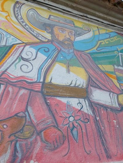 A mural in Municipio Tarija, Bolivia, depicts a man with a beard and a wide-brimmed hat. He is wearing a colorful costume with red, white, blue, yellow, and brown details, and a brown belt. The background includes swirls and geometric shapes in various colors. The mural is painted on a textured wall surface.