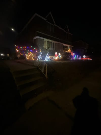 A house is adorned with Christmas lights at night. Strings of multicolored lights are draped along the roof and down the side of the house, with a reindeer made of white lights on the porch. On the porch near the reindeer are Christmas decorations. Steps with a metal railing lead up to the house. A dark shadow appears in the upper right of the picture, obscuring the view.