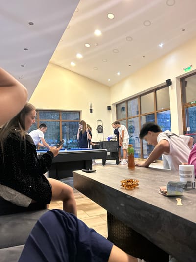 In a brightly lit indoor recreational space, possibly a hotel lobby or entertainment area in Cherna Mesta, Bulgaria, a group of young people are engaged in various activities. In the foreground, a woman with long brown hair, wearing a sparkling black top, is seated and looking at her phone. To her left, a man in a white t-shirt is standing near a pool table, appearing to be playing. Another woman in a dark blue hoodie stands by the pool table, holding a pool cue. Further back, two young men are visible. One, wearing a white t-shirt and shorts, is standing near a dartboard. Another young man, wearing a white t-shirt with a graphic print, is leaning over a table in the mid-ground. The environment features large windows looking out onto an exterior scene, a dartboard, a pool table with balls and cues, and furniture including a long, dark table with a bottle of liquid and some folded bills on it. The ceiling is adorned with numerous recessed lights. An "EXIT" sign is visible on the far right, indicating a doorway.