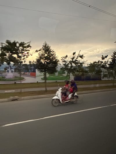 A side view of a white scooter carrying two individuals is captured in motion on a paved road under a cloudy sky, possibly at dusk. The scooter is moving from left to right. The driver, wearing a dark top, is seated in front, and a passenger in a red shirt is behind them. The background features trees and a large billboard or mural displaying architectural renderings and landscaping. The road has a white lane marker. The scene is located in Khan Dangkao, Cambodia.