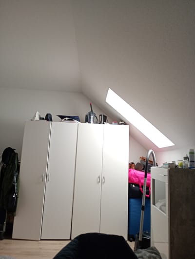 The image depicts an attic room, likely in a residential building located in Osterholz-Scharmbeck, Germany. The room is characterized by a sloped ceiling, illuminated by a large rectangular skylight. In the foreground, a large white wardrobe with four doors is visible, taking up a significant portion of the left side of the frame. To the left of the wardrobe, several dark-colored garments, possibly coats, are hanging on a rack.

On top of the wardrobe, various items are stored, including what appears to be a vacuum cleaner with a red handle, and other indiscernible objects. To the right of the wardrobe, the space is filled with a collection of items, including a bright pink duffel bag, a blue suitcase, and several cardboard boxes. A vacuum cleaner with a grey handle is positioned in front of these items. Further to the right, a white unit with a glass door is visible, with some items stored inside.

The floor is a light-colored wood. The overall impression is that of a cluttered storage space within an attic. No people are present in the image.