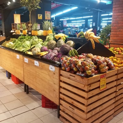 This image captures an indoor produce section within a supermarket located in Kisa Central ward, Kenya. The foreground features a multi-tiered display constructed from light-toned wooden slats, divided into several distinct bins with black trim.
The leftmost bin contains a variety of green leafy vegetables, including cabbages, and a large, light-green gourd. Adjacent bins hold piles of green and purple cabbages, diverse dark-skinned gourds, and bunches of yellow and green bananas. In the background, on a raised black shelf, four pineapples, each with a decorative yellow base, are prominently displayed. The front right section of the display unit contains multiple clear plastic clam-shell packages, each holding several red apples, stacked for sale. A yellow price tag of "299/-" is visible on this section. Behind the apples, a large quantity of oranges or similar citrus fruit is piled.
The wooden display units are supported by black metal legs, with some sections resting on red plastic crates visible beneath. The floor is composed of light-colored square tiles. The overhead environment features a dark ceiling with bright fluorescent strip lighting. No people or animals are visible in the scene. Various unidentifiable signs are partially visible in the distant background.