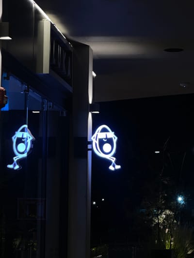 The image depicts the exterior of a commercial establishment at night, located in the city of La Purísima, Mexico. A dark-colored building facade features a prominent, recessed sign above a glass entrance, displaying the word "LUKA" in white block letters. To the right of the entrance, a vertical structural pillar mounts a bright, blue-white neon sign. This sign illustrates a stylized, egg-shaped character with dark sunglasses, an open mouth, and two simple stick-like legs positioned in a relaxed, seated pose. The neon sign is distinctly reflected in the adjacent glass facade to its left, alongside a partial reflection of a red object, likely an internal fire alarm, visible further left within the building. Multiple wall-mounted light fixtures illuminate sections of the dark building exterior. The background to the right is a dark, unlit outdoor area, where sparse, dark vegetation, including tall grasses and slender branches, is faintly discernible, with a distant light source providing minimal illumination.