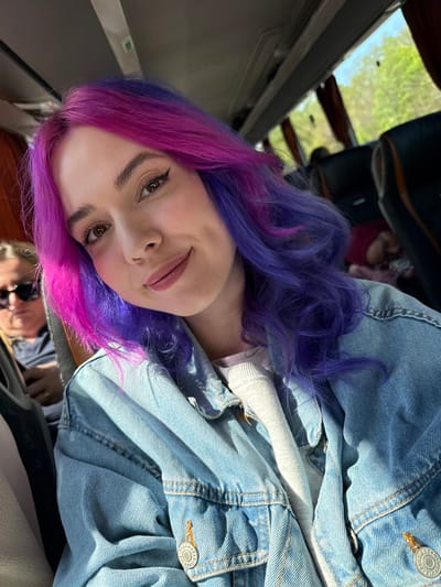 This media file captures a close-up portrait of a young woman in what appears to be the interior of a vehicle, possibly a car or bus. The woman is the central subject, looking directly at the camera with a soft, friendly smile. Her most striking feature is her vibrant, wavy hair, which transitions from a rich purple at the roots to a bright magenta-pink towards the ends. She has brown eyes, subtle eyeliner wings, and light-colored lipstick.

She is dressed in a light blue denim jacket with visible stitching. On her right sleeve, a metal button is partially visible, showing the brand "PULL&BEAR." Beneath her jacket, a lighter-colored garment can be seen. The lighting is bright, suggesting daytime, with strong highlights and shadows on her clothing and hair, indicating a sunny environment.

Through a window in the background, out-of-focus green foliage and a glimpse of a light blue sky are visible, hinting at an outdoor, natural setting, aligning with the location context of Füle, Hungary. The interior of the vehicle shows dark seat backs and window frames. A faint reflection, possibly of a hand holding a phone, can be seen in the lower part of the image. The overall impression is a casual, relaxed moment captured during travel or a stop.