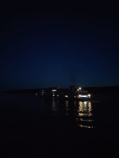 A long, multi-story boat with lit windows moves through dark water at night. The boat is illuminated by its own lights, which reflect on the water. The sky is a deep, dark blue with no visible stars. A dark shoreline is visible in the background. The water appears calm, with the exception of the wake behind the boat. The scene is dark and quiet, suggesting a peaceful and serene environment. The boat appears to be a passenger vessel, possibly a ferry or a cruise ship. The overall impression is one of a tranquil night scene on a body of water.