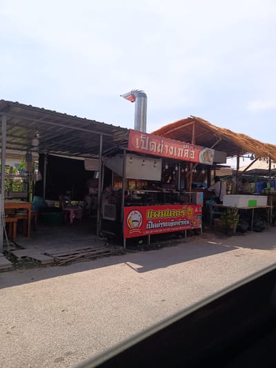 The image shows a street-side food stall in Hua Hin City Municipality, Thailand. The stall is partially covered with corrugated metal roofing and features a thatched roof section. Prominent signage in Thai script advertises "Pet Yang Kleu," (grilled salty duck) and includes a phone number. The stall is equipped with a tall metal exhaust pipe. People can be seen inside the stall. The street is paved.
