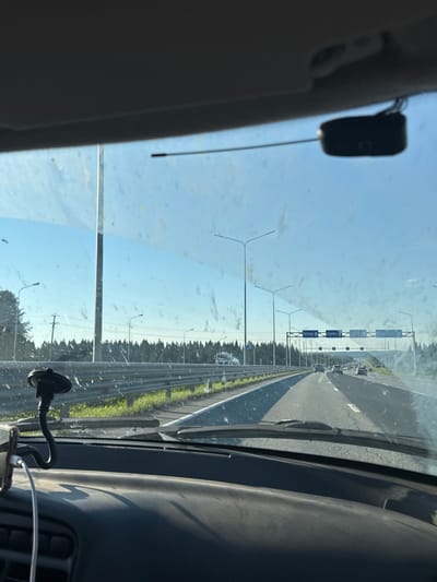 The image shows a view from inside a vehicle driving on a highway. The windshield is dirty with several visible smudges. A black suction cup mount for a device, possibly a phone or dashcam, is attached to the windshield on the driver's side. A thin wire runs from the mount towards the dashboard. The dashboard itself is dark gray and dusty.
Outside the vehicle, the highway stretches ahead, with multiple lanes visible. Roadside barriers are present, with green grass and a line of trees in the background. Several streetlights are positioned along the highway. In the distance, blue road signs with white lettering are suspended over the road. Some of these signs appear to indicate place names in Russian, with "ПЕРМЬ" (Perm) being partially visible on at least two of them. Traffic is light, with a few cars visible in the distance. The sky is clear and blue. The environment appears to be rural or semi-rural, given the presence of trees and open road. The location is identified as being on the highway near Kukushtan, Russia.