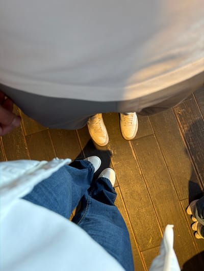 The image presents a high-angle, downward view of the lower extremities of at least three individuals standing on a dark, plank-style wooden floor within an indoor setting in Bretton, United Kingdom.
In the immediate foreground, the legs of one person are visible, clad in wide-leg blue denim jeans and white, flat-soled slip-on shoes. A white fabric, likely part of their upper garment, partially obscures the top of the frame.
Directly opposite, a second individual stands, wearing light grey trousers or shorts and white, lace-up athletic sneakers showing signs of wear. A hand, possibly belonging to this person, is visible on the left, near the hem of the grey garment.
On the far right edge of the frame, the right foot of a third person is partially visible, wearing a light-colored athletic shoe with a distinctive multi-layered sole.
The flooring consists of dark brown wooden planks, arranged linearly, exhibiting visible grain and some surface wear. A warm, yellowish light illuminates the scene, casting subtle shadows. All visible individuals appear stationary.