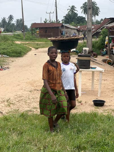 In Ganyingbo, Nigeria, two young girls stand on a dirt road in front of a makeshift market stall. The girl on the left wears a rusty orange tunic and a vibrant green and pink checkered skirt. The girl on the right wears a white t-shirt with a rainbow graphic and dark shorts. Behind them, a table holds various items, including a large cooler labeled "FLORA" and a black plastic basin. In the background, a cluster of corrugated metal roofed buildings line the road, interspersed with palm trees and vegetation. The sky is overcast.