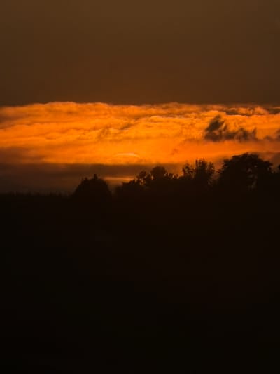 An outdoor scene displays a thick band of orange and yellow clouds against a dark sky, likely at dusk or dawn. The clouds are vertically oriented in the left portion of the image, with darker tones occupying the right side, where indistinct trees or land masses are visible. The overall scene evokes a sense of twilight or impending darkness. There is no information indicating location in the city of Castiglione Tinella, Italy or people, subjects, objects, actions, and interactions.