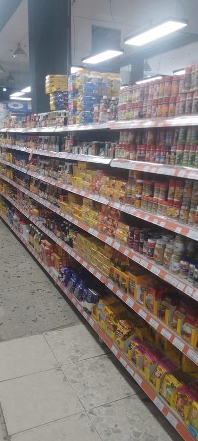 The image depicts an interior view of a supermarket aisle in La Fría, Venezuela. The scene is dominated by multiple levels of commercial shelving, extending diagonally from the lower left towards the upper right, laden with various packaged food products. The shelving units are constructed from metal, appearing white or light gray, with prominent orange strips along the front edge of each shelf displaying pricing information, including "Bs." (Venezuelan Bolívar).

Products visible include numerous clear and colored jars of spices and seasonings, various bottles of sauces and condiments, cereal boxes (some with "Kellogg's" branding), snack bags (including some "Lays" branded products), and packaged goods such as seasoning mixes (some "Maggi" branded) in yellow and other colored boxes and packets. The shelves are densely packed, with products stacked neatly.

The floor consists of light-colored, possibly terrazzo, tiles with dark specks, laid in a grid pattern. Towards the upper left foreground, the flooring transitions to a darker, plainer grey surface, possibly concrete or a different type of tile. Overhead, the ceiling features bright rectangular fluorescent or LED strip lights, along with some spherical pendant lights, providing general illumination. Dark vertical structural columns are visible in the background, contrasting with a lighter ceiling. No people are visible within the frame, and no specific actions or interactions are depicted.