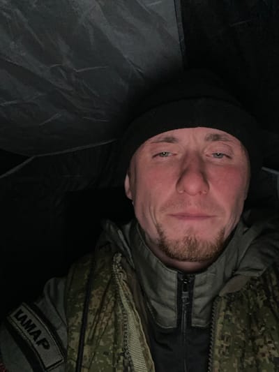 A man wearing a black balaclava, a camouflage jacket, and a patch that reads "KAMAP" is lying down. The environment appears dark and confined, possibly inside a tent or enclosed space. The image is taken from a high angle, looking down at the man's face. The location is unknown and not determinable from the image itself.