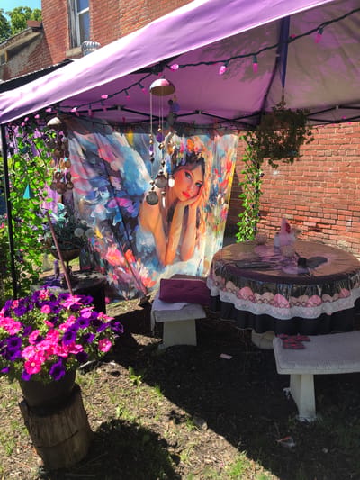 This image depicts an outdoor setup, possibly a decorated seating area in a backyard, under a purple canopy tent. The scene is adorned with various decorative elements. A large fabric hangs as a backdrop, featuring a colorful painting of a woman with wings, reminiscent of a fairy. Pink string lights are draped along the tent's frame, and various hanging ornaments, including beads and shells, are suspended from it.

To the right, a round table is covered with a black tablecloth that has a pink and white scalloped trim. On the table, there are small decorative spheres and a pink object. Adjacent to the table, a concrete bench is visible, with a purple cushion placed on it. Another similar bench is positioned on the left side of the table, also with a cushion.

In the foreground, a large pot overflowing with vibrant pink and purple petunias sits on a weathered tree stump. Green vines with purple accents are entwined on the left side of the setup. The background consists of a brick wall and a glimpse of a building with a window. The ground is covered with dark mulch and some patches of grass. The lighting suggests it is daytime, with shadows cast by the tent and surrounding objects.

The location specified is the city of Severn, Canada.