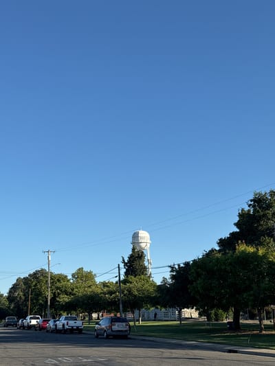 The image displays an outdoor scene, rotated 90 degrees clockwise, depicting a clear day in what appears to be a suburban or semi-rural area. The dominant feature is a large, white spherical water tower located towards the center-right of the frame, partially obscured by trees. The text "RIO L" is visible on the side of the water tower, consistent with the provided location context of Rio Linda.
A vast, unblemished blue sky occupies the upper two-thirds of the image, suggesting clear weather and ample daylight, likely midday or early afternoon given the brightness. Below the sky, a line of lush green trees stretches across the middle ground, casting subtle shadows. To the right, a paved road runs parallel to the line of trees. Several cars, including SUVs and trucks of various colors, are parked along the side of this road. Utility poles with overhead wires are visible, extending horizontally across the image, intersecting with the trees and the sky. A "STOP" message painted on the road surface is partially visible in the bottom right corner. No people or specific activities are observable, presenting a quiet, static street view.