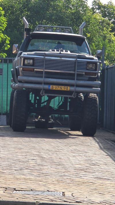 The image shows a custom-built, lifted off-road vehicle, likely a modified Nissan Patrol, parked on a cobblestone surface in 's-Heerenberg, Netherlands. The vehicle is front-facing and dominates the frame, featuring large, knobby tires, a prominent chrome bull bar, and dual exhaust stacks on either side of the hood. A light bar with multiple spotlights is mounted on the roof rack. The vehicle's license plate reads "8-VPN-98". The background consists of lush green trees and a dark metal fence. The scene appears to be outdoors, under daylight, with sunlight casting shadows on the cobblestones. There are no people visible in the image.