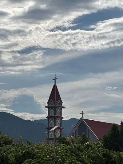 The image depicts a church spire and roof in Suối Tân, Vietnam, under a cloudy sky. The church has a tall, multi-tiered spire topped with a cross, and a sloping red roof. In the foreground, lush green trees obscure the lower parts of the structure. In the background, dark green mountains rise, covered in vegetation. The sky is filled with dramatic, textured clouds, suggesting an overcast or potentially stormy day. There are no people or animals visible in the frame.