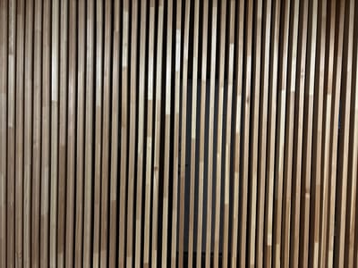 A wall made of vertical wooden slats of light brown color. The slats are evenly spaced, creating a textured pattern. The background behind the slats is dark. The lighting creates shadows on the slats, highlighting their vertical lines and the texture of the wood. There is no information available to determine the location of this wall in Shebarshino, Russia.